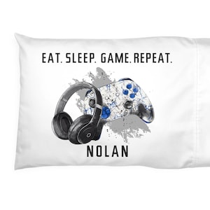 May include: White pillowcase with a graphic design featuring a black headset, a blue and white game controller, and the text "EAT. SLEEP. GAME. REPEAT." and the name "NOLAN".