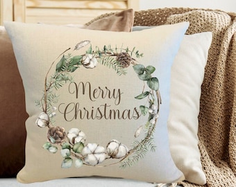 May include: A square beige pillow with a watercolor wreath design and the words "Merry Christmas". The wreath includes cotton, pinecones, and greenery. The pillow is on a gray couch with a tan throw blanket.