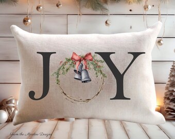 May include: Rectangular pillow with the word "JOY" in large black letters. The "O" is replaced with a watercolor wreath with bells and a red bow. The pillow has a natural or white cover. Text at the top reads "3 SIZE OPTIONS NATURAL OR WHITE COVERS".
