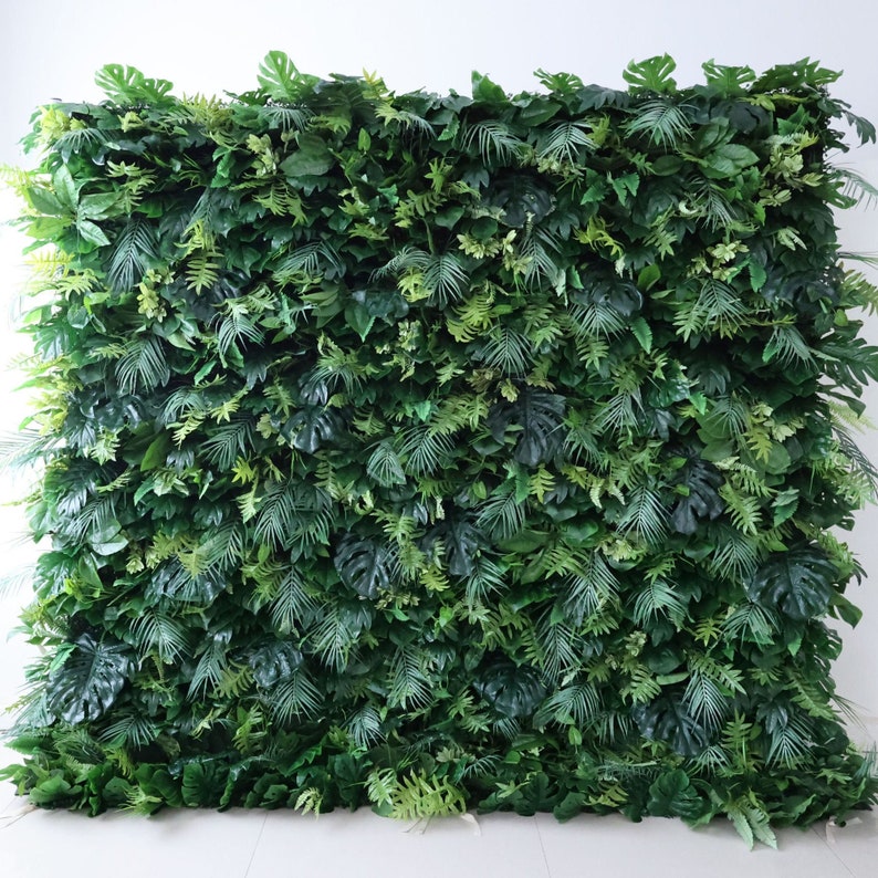 This Valar Flowers lush artificial greenery wall backdrop features a dense, vibrant mix of tropical foliage—including monstera leaves, palm fronds, and ferns—in rich, varied shades of green. The realistic, layered leaves create a fresh, jungle-inspired focal point, perfect for weddings, events, photo booths, and stylish interior decor.