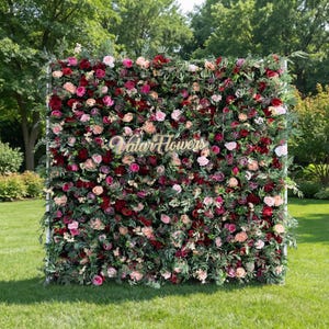 May include: A vibrant floral wall featuring a variety of roses in shades of red, pink, and cream, interspersed with green foliage. The sign "Valar Flowers" is displayed in the center. The arrangement is set against a backdrop of green grass and trees.