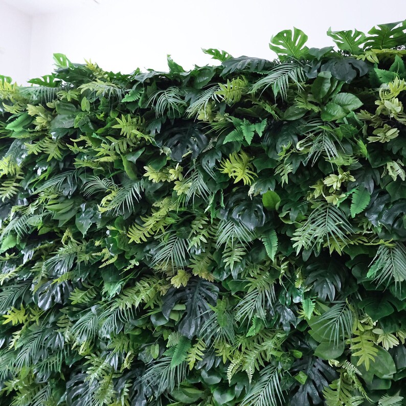 This Valar Flowers artificial tropical greenery wall features a dense, layered mix of lush foliage—including monstera leaves, palm fronds, and ferns—in rich, varied shades of green. The realistic, lifelike leaves create a fresh, jungle-inspired focal point, perfect for weddings, events, photo booths, and stylish interior decor.
