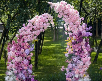 Può includere: Un grande arco floreale caratterizzato da una densa disposizione di rose e ortensie artificiali in varie tonalità di rosa, viola e bianco. I fiori sono raggruppati per formare una struttura curva, creando un vivace arco botanico multicolore.