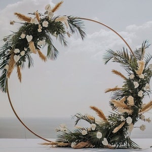 May include: A large, copper-coloured circular wedding arch decorated with white roses, green palm leaves, and dried pampas grass. The arch is set against a backdrop of a cloudy sky and the sea.