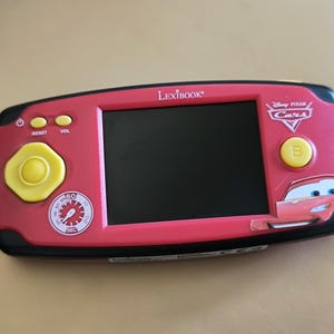 Può includere: Console di gioco elettronica portatile Lexibook rossa con schermo nero, pulsanti gialli e logo Disney Pixar Cars. La console ha una forma rettangolare con angoli arrotondati e bordo nero.