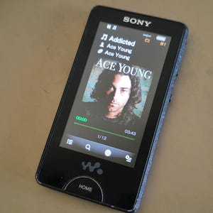 May include: Black Sony digital music player showing the song "Addicted" by Ace Young. The screen displays album art and playback controls. The device features a "HOME" button and a "W" logo.