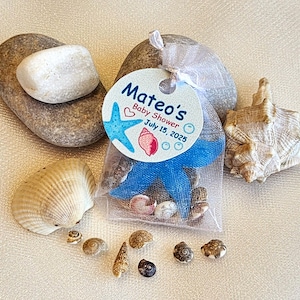 Könnte beinhalten: Ein Babyparty-Gastgeschenk-Set mit einem kleinen Netzbeutel gefüllt mit Muscheln und einem blauen Seestern. Der Beutel ist mit einem Band und einem Etikett mit der Aufschrift "Mateos Baby Shower 15. Juli 2025" gebunden. Das Set enthält verschiedene Muscheln und kleine Steine.