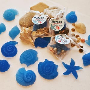 Peut inclure: Divers assortiments de savons en forme de coquillages bleus et de vrais coquillages sont disposés sur une surface blanche. De petits sacs remplis de sable, de coquillages et de pierres sont attachés avec des rubans blancs et étiquetés "Mateo's Baby Shower July 15, 2025".