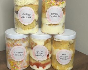 May include: Five glass jars filled with layered cake and frosting. Each jar has a round label with a different cake flavor: Mini Eggs Cake in a Jar, Birthday Cake in a Jar, Old School Cake in a Jar, Jammie Dodger Cake in a Jar, and Lemon Meringue Cake in a Jar.