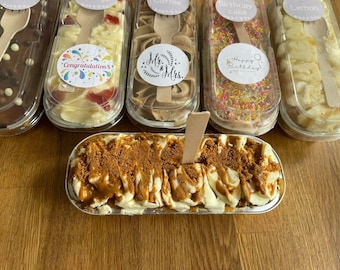 May include: An assortment of individual cakes in clear plastic containers, each with a wooden spoon. Flavors include chocolate, cherry, coffee, birthday cake, and lemon. The bottom cake is topped with a caramel drizzle and cookie crumbs.