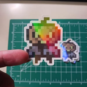 May include: A holographic sticker featuring a pixelated character with a green hat and a blue shirt. The character is standing on a white background.