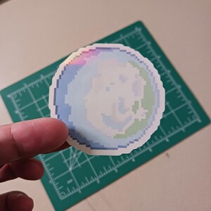 May include: A round sticker with a pixelated design of a smiling face. The sticker features a color gradient of blue, green, and white, with a rainbow iridescent edge. The sticker is on a green cutting mat.