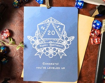 May include: A blue card with a white 20-sided die illustration, surrounded by flowers and a banner that reads "Happy Birthday!"  The card also includes the text "Congrats! You've leveled up".