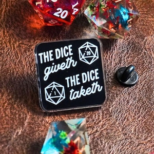 Peut inclure: Épinglette en émail noir avec du texte blanc qui dit "THE DICE giveth THE DICE taketh" avec un graphique de dé à 20 faces à gauche et un graphique de dé à 1 face à droite. L'épingle est entourée de dés colorés.