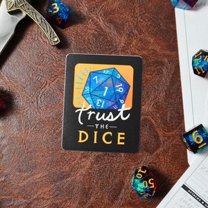 May include: A black and white sticker with a blue and gold iridescent dice with the number 1 showing. The sticker says "Trust the Dice" in a handwritten font.