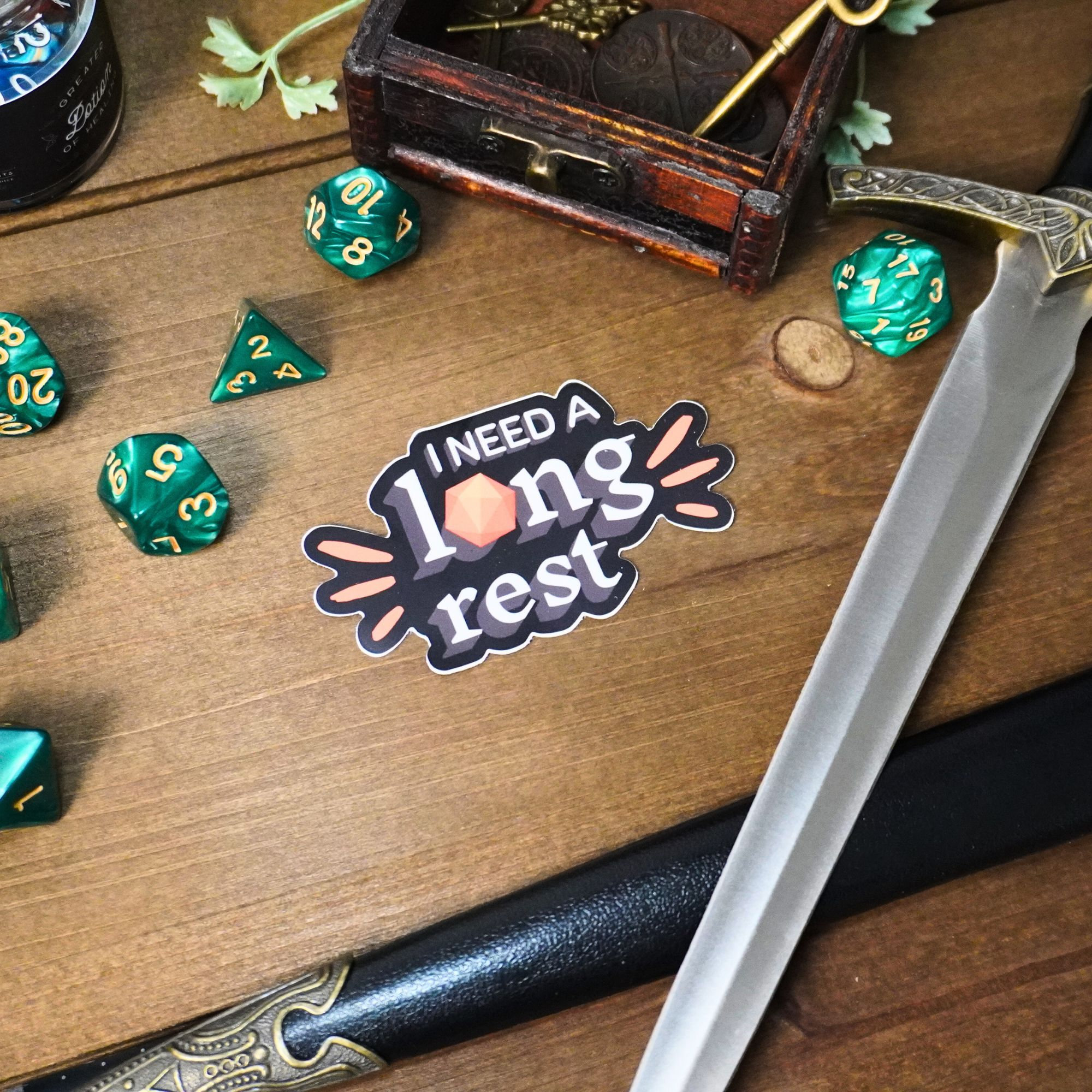 May include: A sticker with the text "I need a long rest" in a black and white font with a pink and orange design. The sticker is on a wooden surface with green dice and a sword.