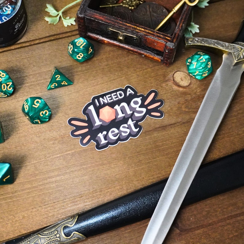 May include: A sticker with the text "I need a long rest" in a black and white font with a pink and orange design. The sticker is on a wooden surface with green dice and a sword.