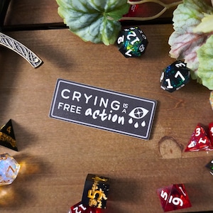 May include: A black and white sticker with the text "Crying is a free action" and a stylized eye with two teardrops. The sticker is on a wooden surface with several colorful dice.