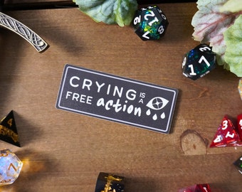 May include: A black and white sticker with the text "Crying is a free action" and a stylized eye with two teardrops. The sticker is on a wooden surface with several colorful dice.