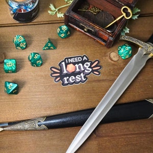 May include: A sticker with the text "I NEED A long rest" in black and white with a peach-colored die in the middle. The sticker is on a wooden surface with green dice, a sword, and a wooden box with a key.