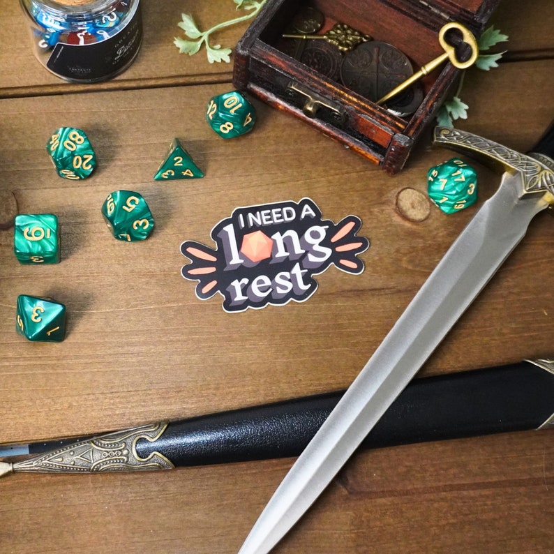 May include: A sticker with the text "I NEED A long rest" in black and white with a peach-colored die in the middle. The sticker is on a wooden surface with green dice, a sword, and a wooden box with a key.