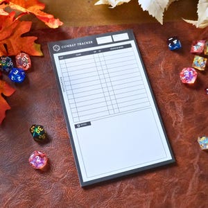 May include: A black and white Combat Tracker notepad with a grid for tracking game stats. The notepad is surrounded by colorful dice and autumn leaves on a brown surface.