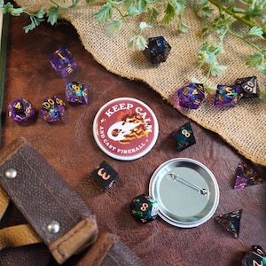 Pode incluir: Um botão vermelho e branco com o texto "Keep Calm and Cast Fireball" rodeado por dados roxos, dourados e azuis.