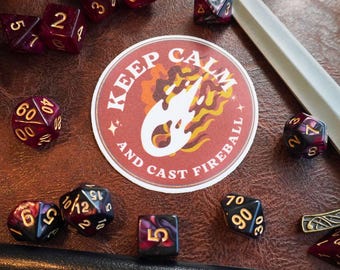 May include: A white sticker with red, orange, and yellow flames and the text "Keep Calm and Cast Fireball" on a brown leather surface with red and black dice.