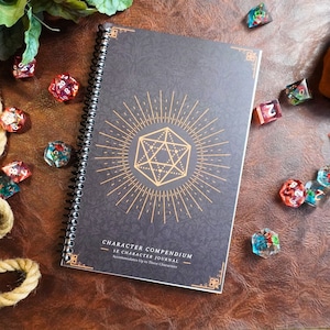 May include: A spiral-bound Character Compendium journal with a gold geometric design on a dark gray cover. The journal is surrounded by colorful polyhedral dice on a brown leather surface. Text on the cover reads "CHARACTER COMPENDIUM 5E CHARACTER JOURNAL Accommodates Up to Three Characters."