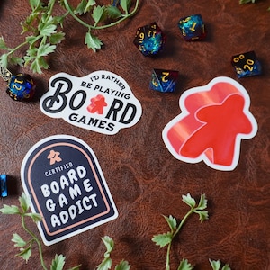 May include: Two stickers with a board game theme. One sticker is white with black text that says "I'd rather be playing board games". The other sticker is white with a black arch and the text "Certified Board Game Addict".