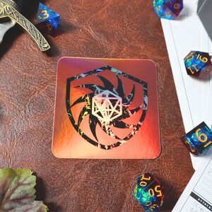 May include: A holographic sticker with a black and white design of a shield with a swirling pattern and a white d20 die in the center. The sticker is on a brown leather surface with dice scattered around it.