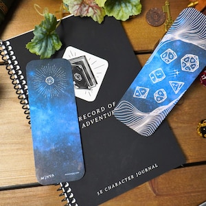 May include: Two blue and white galaxy themed bookmarks with dice and a starburst design. The bookmarks are laying on top of a black journal with the words "Record of Adventure" and "5E Character Journal".