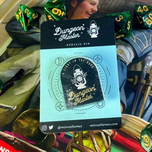 May include: An acrylic pin with the words "Dungeon Master" and a lantern design. The pin is on a light blue backing card with the text "A light in the dark." Green and gold dice are scattered around the pin.