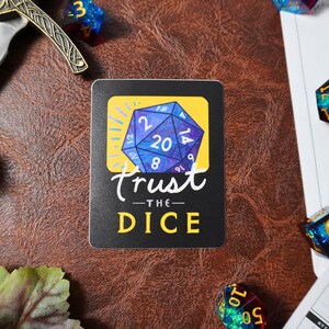 May include: A black and white sticker with a blue and yellow dice with the numbers 2, 8, 12, 14, 20, and 9. The text "Trust the Dice" is written in white cursive lettering.