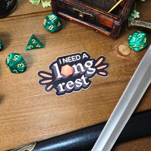 May include: A sticker with the text "I need a long rest" in a retro style font. The sticker features a stylized orange d20 dice and a black background.
