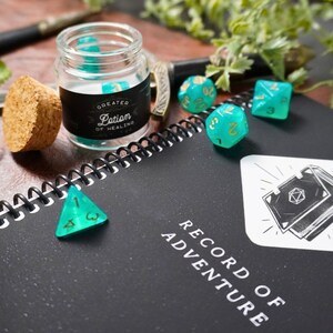 May include: A black notebook with the words "Record of Adventure" on the cover.  There are several green and gold dice on the notebook, and a small glass jar with a black label that reads "Greater Lotion of Healing".