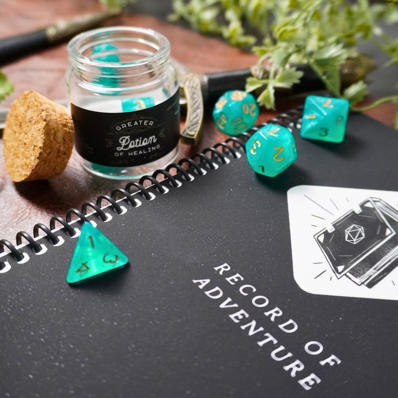 May include: A black notebook with the words "Record of Adventure" on the cover.  There are several green and gold dice on the notebook, and a small glass jar with a black label that reads "Greater Lotion of Healing".