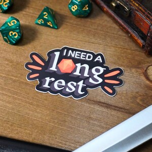 May include: A black and white sticker with the text "I need a long rest" and a red 20-sided die. The sticker has orange accents.
