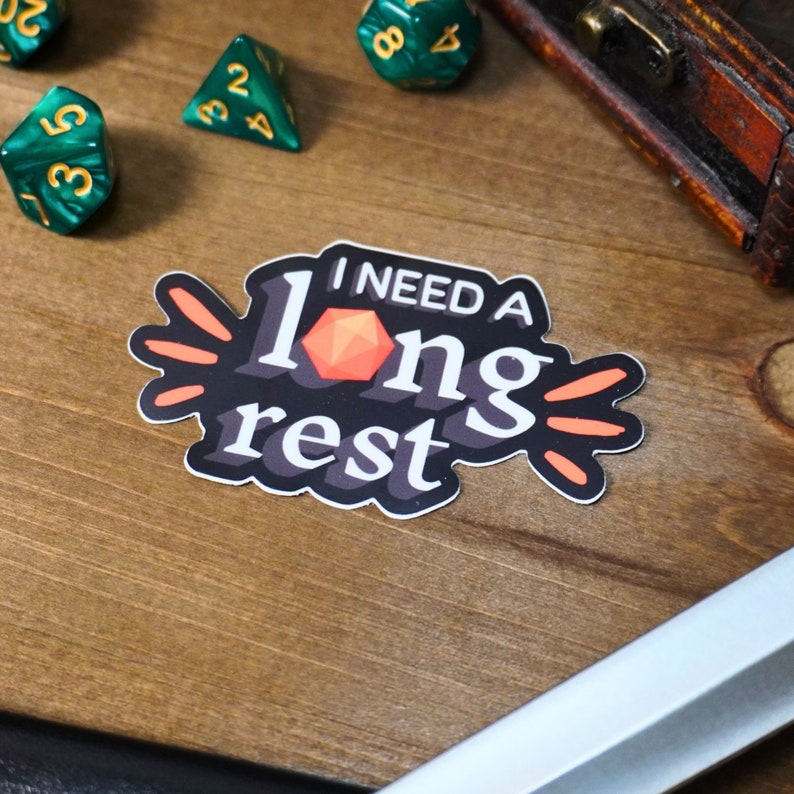 May include: A black and white sticker with the text "I need a long rest" and a red 20-sided die. The sticker has orange accents.