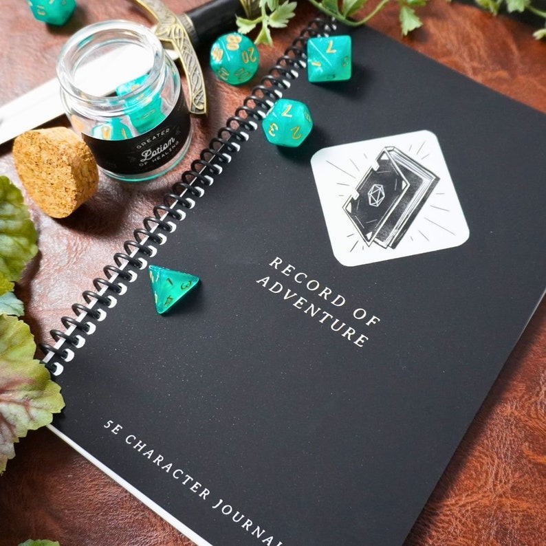 May include: A black notebook with a gold spiral binding and the text "Record of Adventure" and "Character Journal". The notebook is surrounded by teal and green dice.