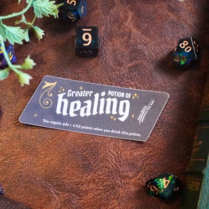 May include: A black and white sticker with gold lettering that reads "Greater Potion of Healing". The sticker is on a brown leather surface with dice scattered around it. The text on the sticker reads "You regain 4d4 + 4 hit points when you drink this potion."