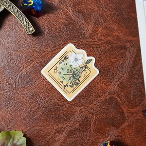 May include: A sticker featuring a green 20-sided die with a white flower and green leaves on a yellow background. The die is showing the number 20.