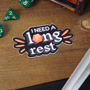 May include: A black and orange sticker with the text "I need a long rest" and a red 20-sided die. The sticker is on a wooden surface.