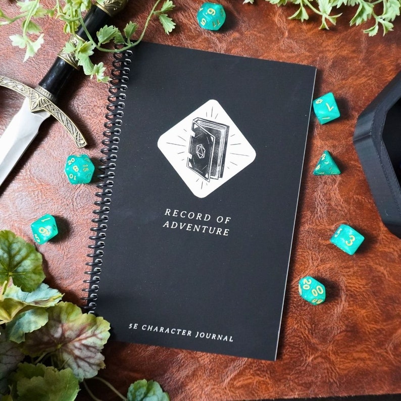 May include: A black spiral bound journal with a white illustration of a closed book on the cover. The text "Record of Adventure" and "5e Character Journal" are printed on the cover. The journal is surrounded by green dice.