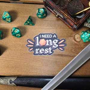 May include: A sticker with the text "I need a long rest" in a playful font. The sticker features a stylized orange d20 dice and a peach background. The sticker is on a wooden surface with green dice and a sword.