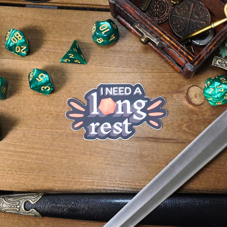 May include: A sticker with the text "I need a long rest" in a playful font. The sticker features a stylized orange d20 dice and a peach background. The sticker is on a wooden surface with green dice and a sword.