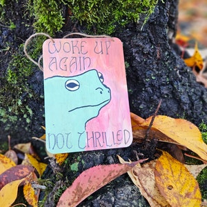 Può includere: Un cartello di legno con un'illustrazione di rana verde e il testo "Woke up again not thrilled". Il cartello è appeso a un ramo d'albero con muschio e foglie autunnali.