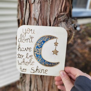 Puede incluir: Un letrero de madera con una luna creciente azul y dorada y estrellas. El letrero dice "You don't have to be whole to shine."