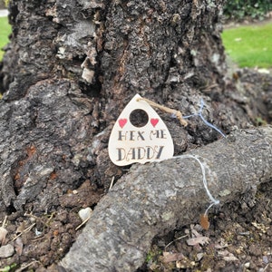Puede incluir: Un letrero de madera con forma de fantasma con un agujero en el medio y una cuerda atada. El letrero dice "Hex Me Daddy" con dos corazones rojos encima del texto.