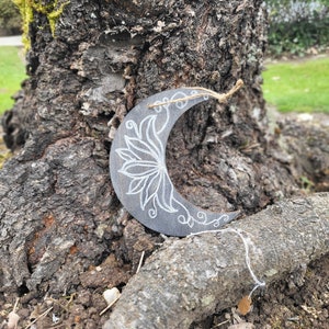 Puede incluir: Un adorno de madera en forma de media luna con un diseño floral blanco pintado sobre él. El adorno está colgado de una rama de árbol.